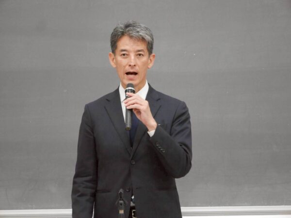 Professional speaker in a dark suit delivering a talk, holding a handheld microphone in front of a chalkboard.