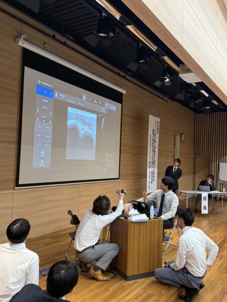 Lecture room with a projected screen displaying software UI; a presenter in a suit stands by a banner while attendees observe and discuss.